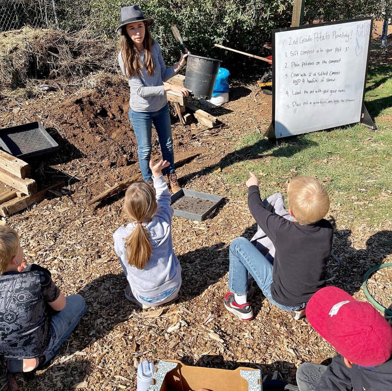 Children raise their hands while a woman holds a planting pot beside a board with potato-planting instructions.