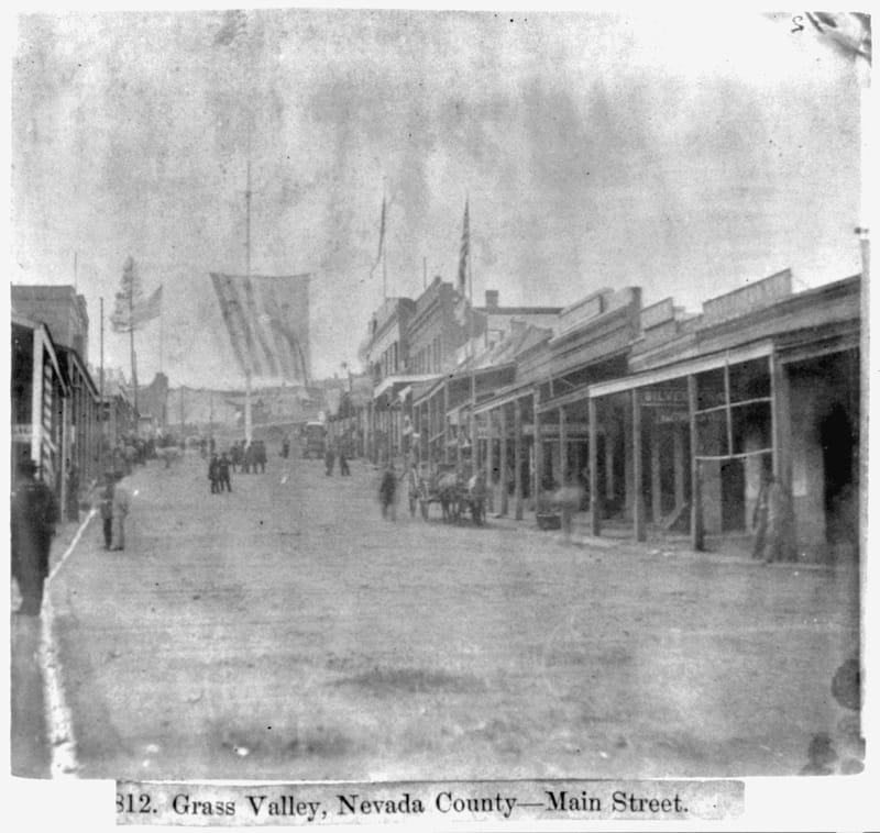 Historic photograph of Grass Valley’s unpaved Main Street, lined with storefronts and covered sidewalks, with people and horses in the street.